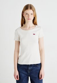 Krém színű, testhezálló póló kerek nyakkivágással és rövid ujjakkal, a mellkasán egy kis piros Levi's logóval. Kék farmernadrággal viselve.