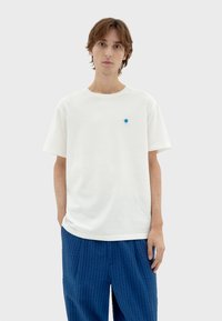 Witte katoenen t-shirt met korte mouwen, voorzien van een klein blauw cirkelvormig logo op de borst. Het model draagt gestructureerde blauwe broek.