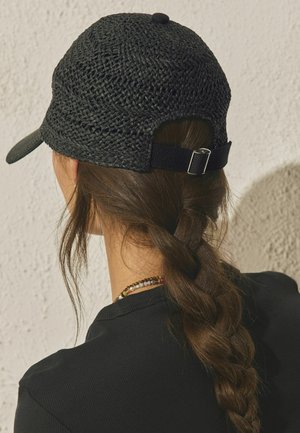 Woman with long brown braid wearing a black woven cap with adjustable strap, black shirt, and a beaded necklace, facing textured light wall.