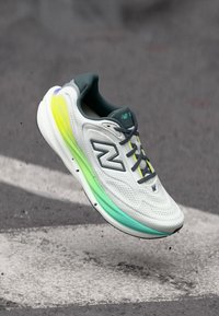 Λευκό και γκρι παπούτσι τρεξίματος New Balance με νέον κίτρινες και πράσινες λεπτομέρειες, αιωρούμενο πάνω από την άσφαλτο.