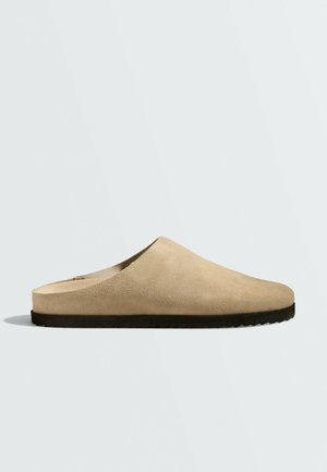 Chaussure beige à enfiler avec dessus en daim souple et semelle en caoutchouc noire, conçue pour un port facile et un confort en intérieur comme en extérieur.