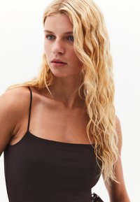 Top nero senza maniche con scollatura quadrata, texture liscia e design minimalista, indossato da un modello con capelli biondi e mossi.