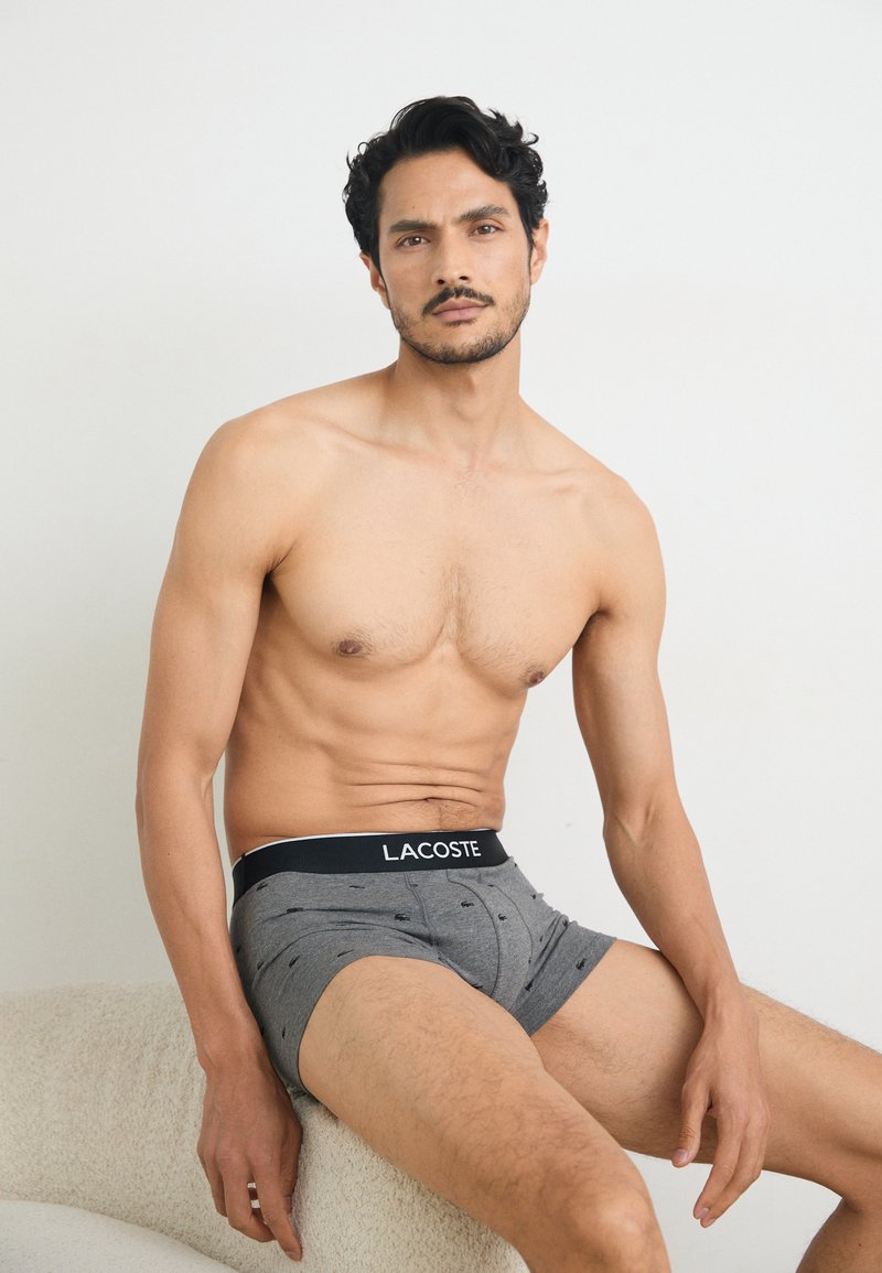 Gray boxer briefs with a black waistband featuring the "LACOSTE" logo; subtle pattern of small motifs on the fabric.