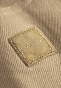 Beige stof med en syet firkantet lap med det prægnede ord "GABBA" i midten, der fremhæver tekstur og detaljer.
