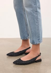 Melnas slingback dzīvokļi, izgatavoti no teksturēta auduma ar zebru joslu rakstu, ar norādītu pirkstu un zemu bloku papēdi. Valkā ar gaiši ziliem džinsiem.