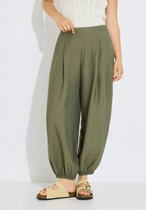 Mujer con pantalones sueltos verdes oliva con puños elásticos y sandalias beige de punta abierta, de pie frente a un fondo blanco liso.