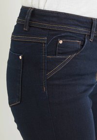 Jeans in denim blu scuro con cuciture arancioni a contrasto, tasca frontale e design aderente. Texture liscia e cintura standard.