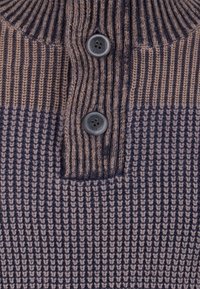 Närbild av stickad tröjfabric i brun och marinblå med vertikala ribbor och två mörkbruna knappar på en knappslå.