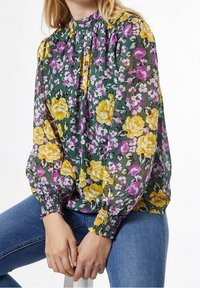 Blusa floreale in tessuto verde trasparente con rose gialle e fiori viola, con colletto e polsini smock. Abbinata a jeans blu.