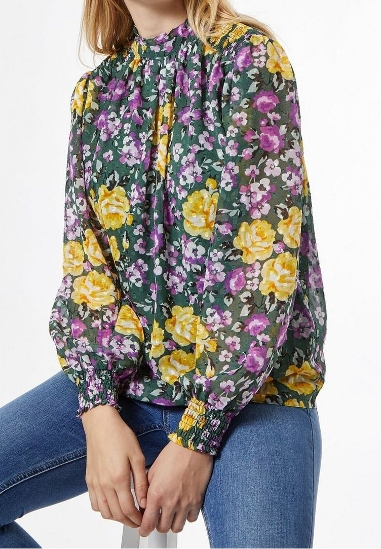 Blusa floreale in tessuto verde trasparente con rose gialle e fiori viola, con colletto e polsini smock. Abbinata a jeans blu.