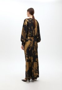 Lang jurk in zwart en goud met een tie-dye patroon, gemaakt van een zijdezachte stof. Bevat lange mouwen en een relaxed fit met knoopaccenten.
