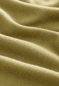 Blødt, struktureret stof i lys olivengrøn, med blide folder og en glat overflade, der fremhæver dens strikkemønster og elasticitet.