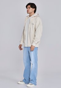 Hellbeige Zip-Hoodie mit weiter Passform und Kängurutasche, getragen über einem weißen Hemd; kombiniert mit hellblauen Jeans und einfachen Sneakers.