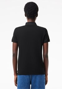 Personne aux cheveux courts et bouclés portant un polo noir uni à manches courtes et un jean en denim bleu, vue de dos.
