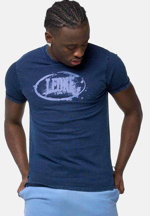 Jonge man met gevlochten haar, een donkerblauw "Leone 1947" T-shirt en lichtblauwe broek dragend, kijkt naar beneden met handen in de zakken.