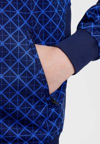 Veste à motifs bleus avec un design géométrique, poche zippée sur le côté et poignets côtelés. Texture lisse avec un fond bleu foncé.