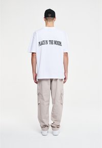 Witte t-shirt met grote zwarte tekst op de achterkant met de tekst "PLAATS IN HET BOS." Gecombineerd met beige cargobroek en witte sneakers.