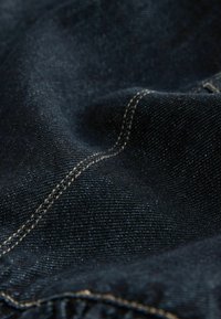 Gros plan d’un tissu en denim bleu foncé montrant un tissage texturé et une double surpiqûre beige le long d’une couture.