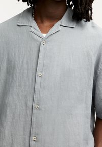 Camisa de lino gris claro de manga corta con cuello de muesca, que cuenta con cinco botones en el frente y un corte relajado. Textura suave.