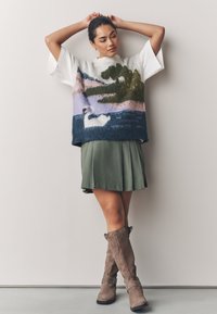 Pull tricoté multicolore avec motif paysager sur une chemise blanche, assorti à une jupe plissée verte et des bottes hautes marron.