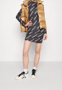 Veste matelassée dorée sur une robe noire à manches longues avec le logo blanc "adidas" répété. Associée à des baskets blanches avec des rayures noires.