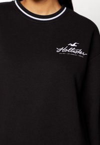Czarna bluza z okrągłym dekoltem, z białym prążkowanym kołnierzykiem oraz haftowanym logo "Hollister" z przodu.