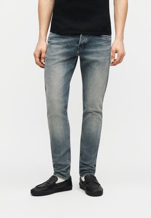 Muž oblečený do vybledlých modrých slim fit džínsov, čiernych topánok a čiernej košele, stojaci pred svetlosivým pozadím.