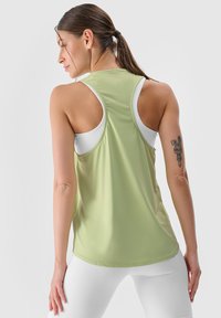 Top atletico senza maniche di colore verde chiaro con tessuto testurizzato. Presenta un collo a barca e ampi fori per le braccia, indossato sopra un reggiseno sportivo bianco.