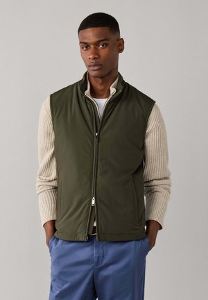Jonge man die een groene zip-up bodywarmer draagt over een beige trui en blauwe broek, staand met handen in de zakken tegen een effen achtergrond.