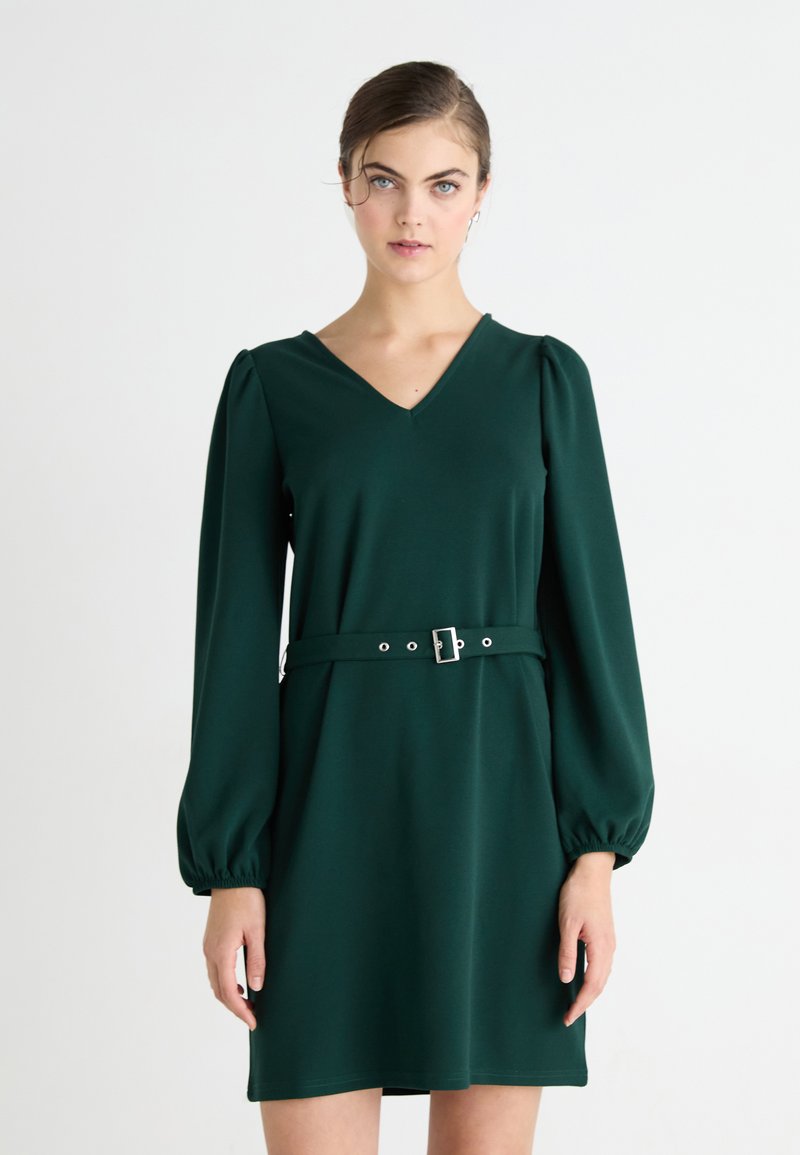Zelené šaty vyrobené z hladkej tkaniny, s výstrihom do V, napufkanými dlhými rukávmi a opaskom so striebornou prackou. Jednoduchý a priliehavý dizajn.