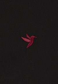 Tejido negro con un colibrí bordado en rojo con las alas extendidas, textura detallada y definición clara contra el fondo.