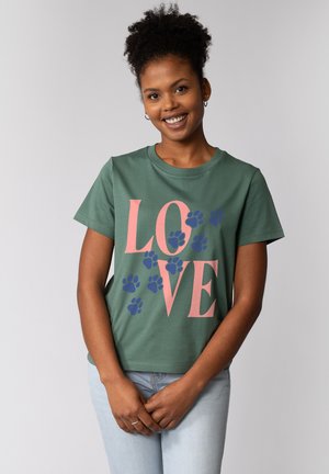 Lächelnde Frau mit lockigem Haar, die ein grünes T-Shirt mit pinkem "LOVE"-Schriftzug und blauen Pfotenabdrücken trägt, und vor einem einfarbigen Hintergrund steht.