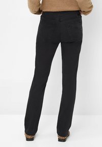 Person, der schwarze gerade geschnittene Jeans und Schuhe mit Leopardenmuster trägt, steht mit dem Rücken zu einer schlichten weißen Wand.