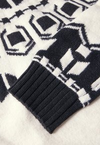 Sort og hvid strikket sweaterærme med geometriske mønstre, der hviler på en baggrund af matchende mønstret stof.