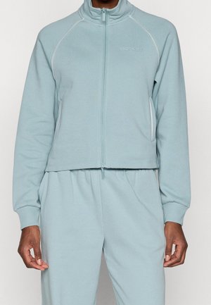 Homme portant une veste zippée bleu clair et un pantalon assorti avec le logo « ONLY PLAY » sur la poitrine, debout devant un fond uni.