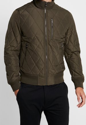 Mann trägt olivgrüne gesteppte Jacke mit Reißverschlusstaschen und schwarze Hose, steht mit entspannten Händen an den Seiten.