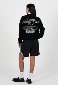 Czarny sweter z dzianiny z napisem "IN A RUSH Racing Club BERLIN" i grafiką samochodu na plecach, zestawiony z czarnymi szortami i czarnymi butami.