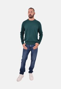 Grüner Strickpullover mit Rundhalsausschnitt, kombiniert mit blauen Jeans und weißen Sneakern. Der Pullover hat ein kleines Logo auf der Brust und gerippte Bündchen.