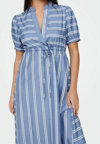 Ljusblå omlottklänning med korta volangärmar, som har vertikala vita ränder och ett knyt som detalj i midjan. Tillverkad av mjukt tyg med en slät yta.