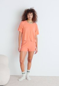 Korallfärgad bomullsloungewear-set med en kortärmad, kort topp och matchande shorts. Kombinerat med knähöga vita strumpor med gröna ränder.