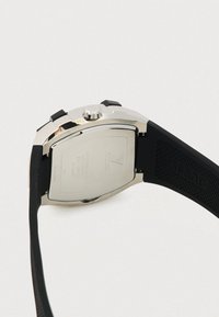 Dos de montre en acier inoxydable argenté avec un bracelet en caoutchouc noir, présentant une forme circulaire, une surface lisse et des marques gravées.