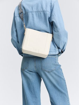 Sac à main en cuir couleur crème de forme rectangulaire, avec une bandoulière à motifs et une surface texturée. Porté sur une tenue en denim.
