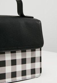 Sac à main en cuir noir avec un panneau avant en tissu à carreaux noir, blanc et gris, et une anse supérieure.