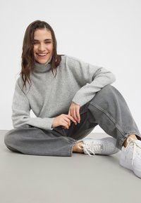Světle šedý oversized svetr s vysokým límcem, žebrovanými manžetami, v kombinaci s volnými šedými džíny a bílými teniskami. Posed v neutrálním pozadí.