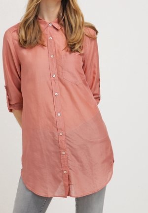 Woman wearing a long, loose, buttoned rose-colored shirt with rolled-up sleeves and front chest pocket, paired with gray pants.