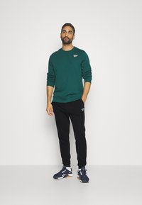 Sweatshirt vert avec un col rond, joggeurs noirs et baskets navy. Présente un petit logo sur le sweatshirt et les joggeurs.