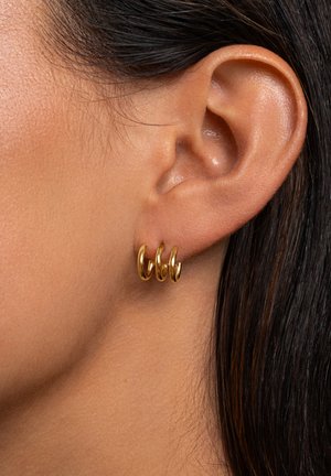 Gros plan sur une oreille gauche ornée de trois petites boucles d'oreilles en or lisses, avec des cheveux raides et foncés visibles sur le côté droit.