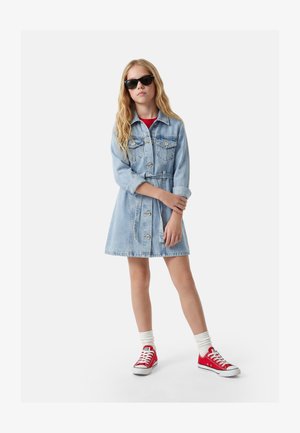 Chica con cabello largo y rubio que lleva gafas de sol negras, un vestido ligero de mezclilla con cinturón, calcetines blancos y deportivas rojas, de pie con los brazos cruzados.