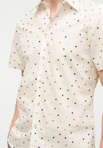 Κοντομάνικο κρεμ πουκάμισο με πολύχρωμο μικρό τετράγωνο σχέδιο, κουμπωμένο μπροστά, φοράει ένας άνθρωπος με τα χέρια στις τσέπες.