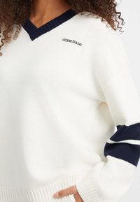 Fehér pulóver tengerészkék V-nyakkal, "GUESS JEANS" logóval és tengerészkék csíkkal a ujjakon. Puha tapintású, bordázott alsó szegéllyel.